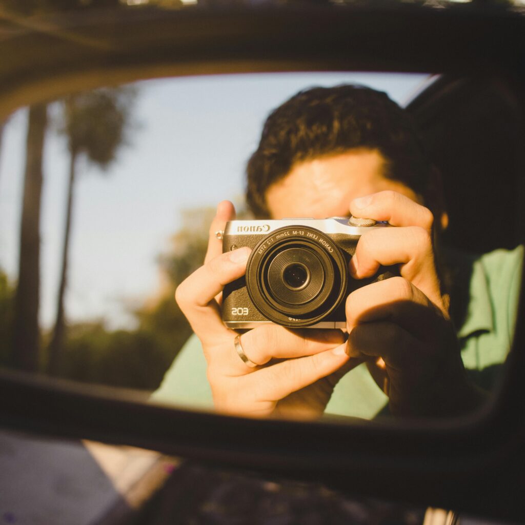 A person sitting in a car taking pictures with a retro camera through the window for the blog "The Small Moments We Keep Becoming" by Dhrruv Tokas.