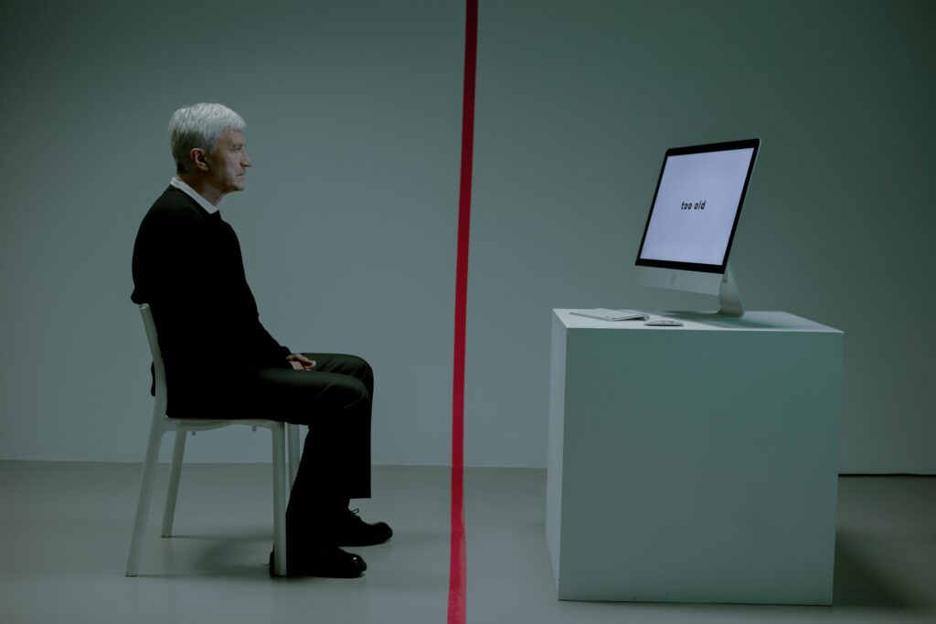 A senior man sits in a dimly lit room, his face etched with contemplation as he stares at a computer screen displaying the words 'too old' in bold text. This image illustrates the external pressures of ageism and the internal struggle of self-definition, from the blog "When You Decide Who You Are Too Soon" by Dhrruv Tokas.