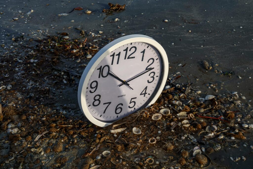 An evocative shot of a vintage clock half-buried in the wet sand of a shoreline, surrounded by scattered seashells as waves wash nearby. The image symbolizes the futility of waiting for the perfect moment and the erosion of "the right time" by the tide of procrastination; from the blog "The Myth of the Right Time" by Dhrruv Tokas.
