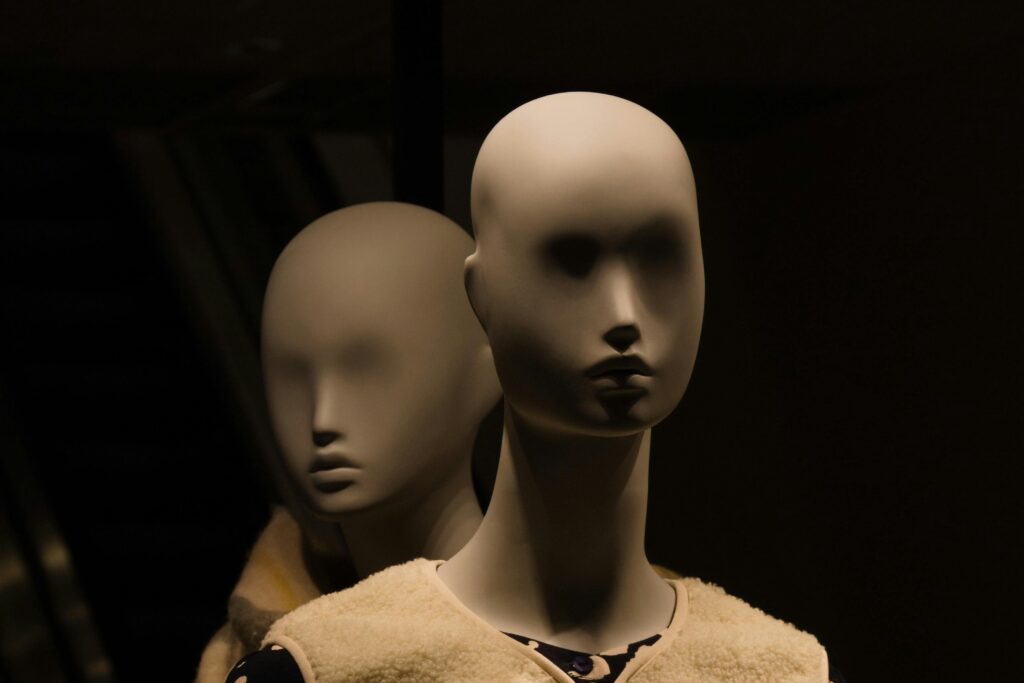 A dramatic close-up of two featureless mannequins standing close together in dim, atmospheric lighting. The shadows cast across their blank faces create a mysterious, eerie ambiance, visually capturing the unsettling feeling of a human identity being cloned into a hollow, artificial double. The moody setting highlights the core themes of digital duplication and the loss of personal sovereignty explored in the blog "Who Owns Your Voice" by Dhrruv Tokas.