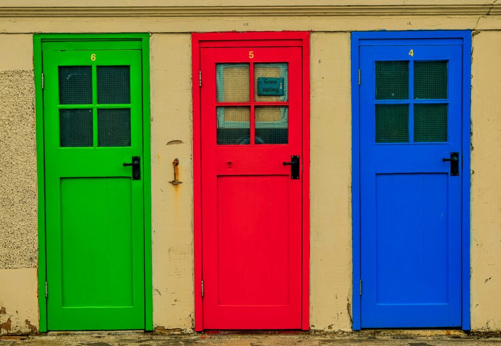Three distinct, brightly colored wooden doors—one green, one red, and one blue—standing side by side against a neutral wall. The stark choices symbolize the hidden pressure of modern decision-making and the quiet strength found in committing to a path, from the blog "The Art of Closing Doors" by Dhrruv Tokas.