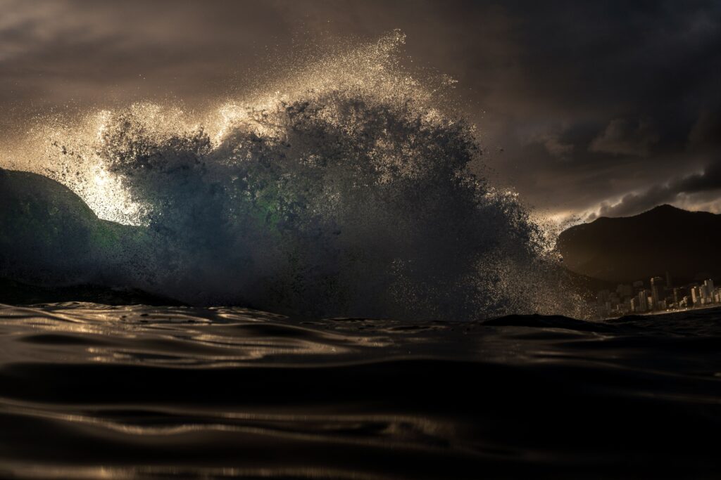 A dramatic, powerful ocean wave crashing violently against a coastal cityscape, illuminated by the fiery light of a deep sunset. The juxtaposition of the volatile, rising tide against the permanent city buildings beautifully mirrors how sudden emotional storms crash into our daily lives, illustrating the theme of emotional stewardship in the essay "Anger as Weather" by Dhrruv Tokas.