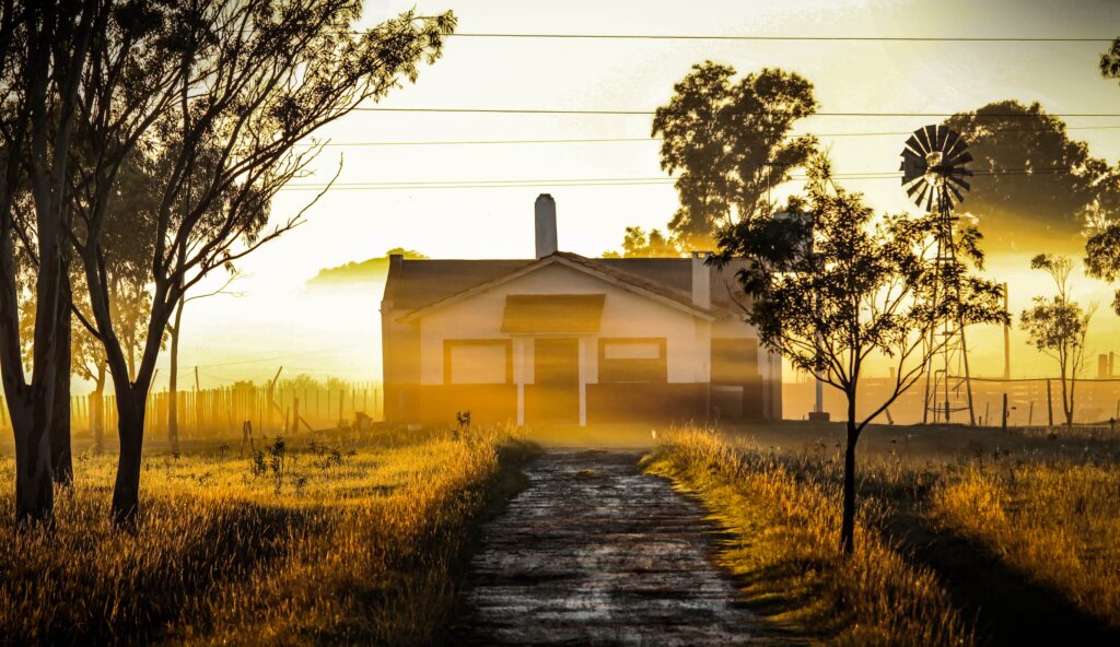 A wide shot of a serene countryside scene featuring a rustic house and a traditional windmill standing in a misty field at sunrise. The soft morning light breaking through the fog beautifully symbolizes dawn, memory, and the distant, evolving nature of home, highlighting the core themes of the blog "Home as a Moving Target" by Dhrruv Tokas.